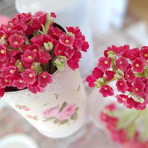 May include: A decorative arrangement of vibrant red flowers with yellow centers and green accents. The flowers are arranged in a vintage-style tin can adorned with a floral pattern and lace trim, creating a charming, rustic aesthetic.