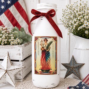 May include: A white painted glass bottle with a red glitter bow and a vintage patriotic label. The label features a woman in a dress with the American flag design. The bottle is decorated with a red glitter bow and is surrounded by patriotic decor.