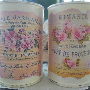 Op de afbeelding: Twee vintage metalen blikken met bloemmotieven. De blik links heeft de tekst "ELLE JARDINIERE 2. Rue du Pont Neuf, PARIS CARTE POSTALE Ce site est exclusivement reserve a l'adherent" en de blik rechts heeft de tekst "ROMANCE PAR ESSENCE CONCENTREE ROSE DE PROVENCE".
