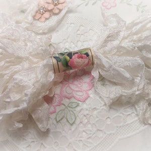 May include: A white lace ribbon spool with a floral design. The spool is wrapped in a white lace ribbon and has a floral design on the front. The spool is sitting on a white lace tablecloth.