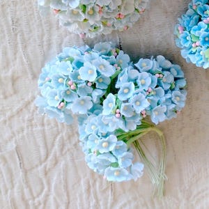 Pu&ograve; includere: Un grappolo di fiori artificiali azzurri con piccoli centri gialli e minuscoli accenti rosa, fissati su sottili steli di filo metallico verde. I petali arrotondati sono disposti in gruppi densi e sferici, creando una composizione floreale.