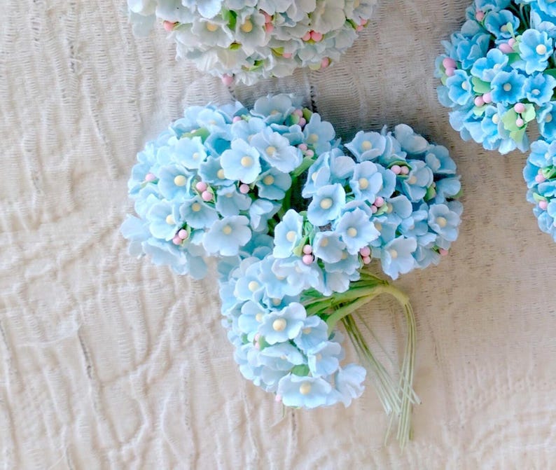 Pu&ograve; includere: Un grappolo di fiori artificiali azzurri con piccoli centri gialli e minuscoli accenti rosa, fissati su sottili steli di filo metallico verde. I petali arrotondati sono disposti in gruppi densi e sferici, creando una composizione floreale.