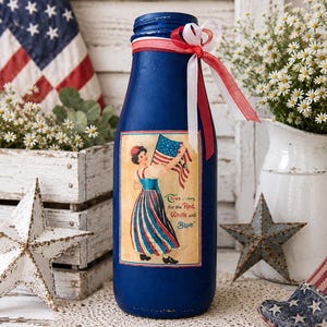 May include: A blue decorative bottle with a patriotic theme. It features a vintage-style label with a woman holding an American flag and the text "Three colors for the Red, White and Blue." The bottle is adorned with red and white ribbons.