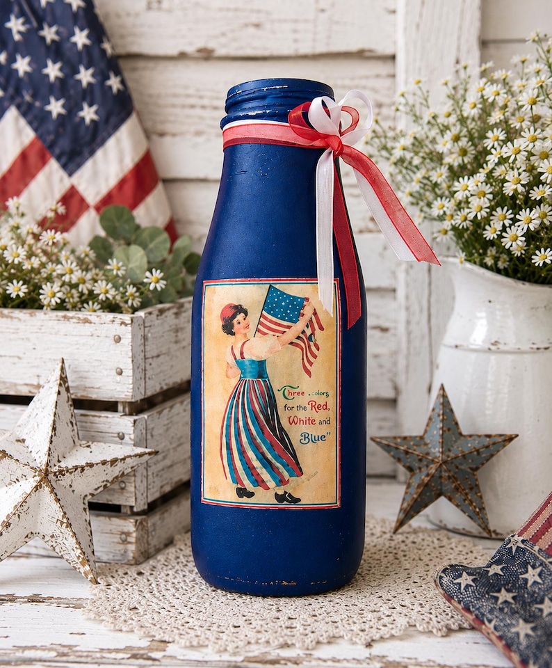 May include: A blue decorative bottle with a patriotic theme. It features a vintage-style label with a woman holding an American flag and the text "Three colors for the Red, White and Blue." The bottle is adorned with red and white ribbons.