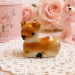 May include: A small ceramic figurine of a fawn in a resting position. The fawn has a glossy brown coat with white spots on its back, black eyes, a black nose, and black hooves. It is positioned on a white crocheted lace doily.