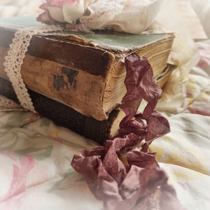 May include: Two vintage books with worn leather covers are stacked on top of each other. A delicate lace ribbon is tied around the top book, and a pink flower is placed on top of the ribbon. A piece of burgundy fabric is draped over the books and hangs down the side.