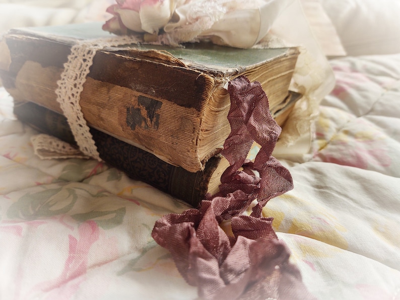 May include: Two vintage books with worn leather covers are stacked on top of each other. A delicate lace ribbon is tied around the top book, and a pink flower is placed on top of the ribbon. A piece of burgundy fabric is draped over the books and hangs down the side.