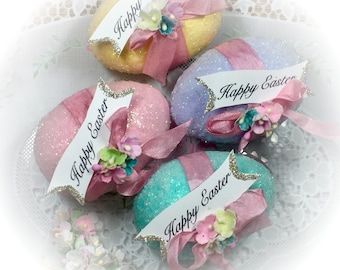May include: Four glittery pastel Easter eggs decorated with pink ribbon and floral accents. Each egg has a white banner with the text "Happy Easter" in black.