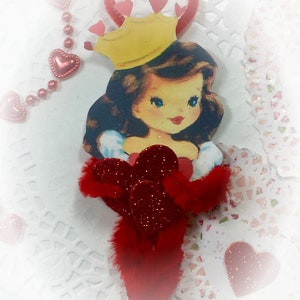May include: A red and white Valentine's Day ornament featuring a cartoon princess wearing a yellow crown and a red dress. The ornament is decorated with red glitter and hearts.