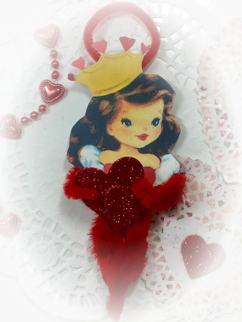 May include: A red and white Valentine's Day ornament featuring a cartoon princess wearing a yellow crown and a red dress. The ornament is decorated with red glitter and hearts.