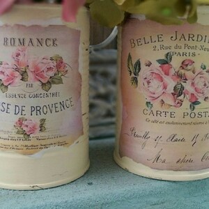 Op de afbeelding: Twee vintage metalen kannen met bloemmotieven en Franse tekst. De kan links zegt "RUMANCE PAR ESSENCE CONCENTREE ROSE DE PROVENCE". De kan rechts zegt "BELLE JARDINIERE 2. Rue du Pont Neuf PARIS CARTE POSTALE".