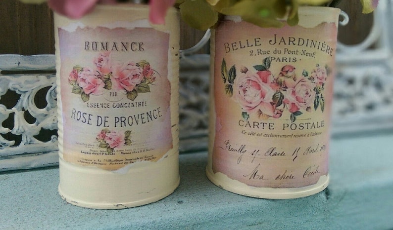 Op de afbeelding: Twee vintage metalen kannen met bloemmotieven en Franse tekst. De kan links zegt "RUMANCE PAR ESSENCE CONCENTREE ROSE DE PROVENCE". De kan rechts zegt "BELLE JARDINIERE 2. Rue du Pont Neuf PARIS CARTE POSTALE".