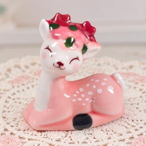 May include: A small ceramic deer figurine in a resting position. The deer is primarily light pink with a white chest and face, featuring white spots on its back, a dark red antler crown with green leaf accents, and a black hoof visible on the side.