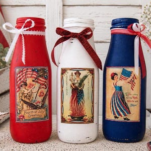 May include: Three decorative glass bottles painted red, white, and blue, with patriotic labels and ribbon accents. Each bottle features a vintage-style image of a woman with an American flag or holding candles. The bottles are decorated for the Fourth of July.