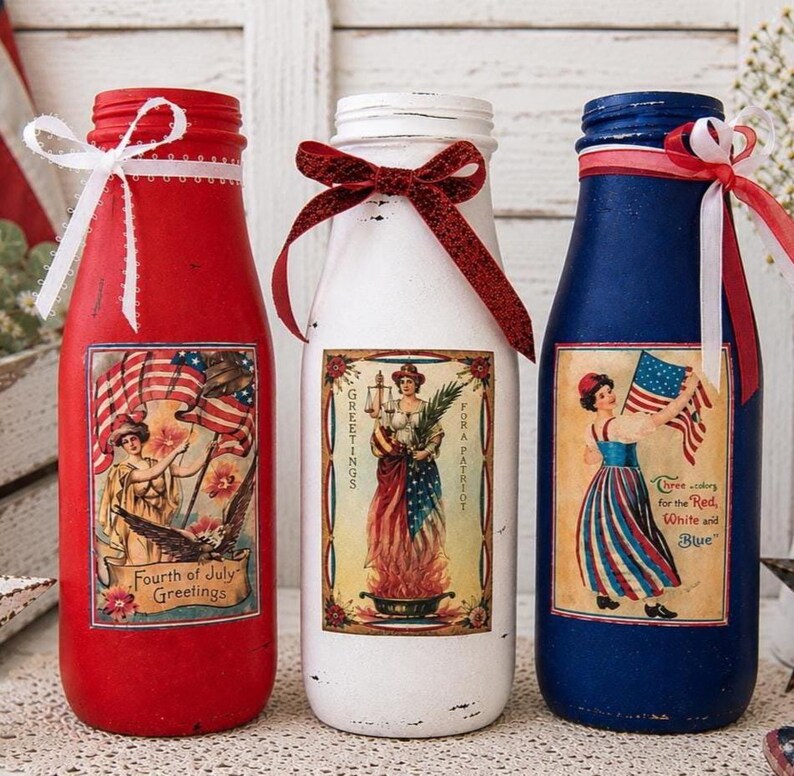 May include: Three decorative glass bottles painted red, white, and blue, with patriotic labels and ribbon accents. Each bottle features a vintage-style image of a woman with an American flag or holding candles. The bottles are decorated for the Fourth of July.