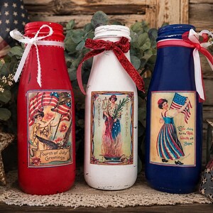 May include: Three painted glass bottles in red, white, and blue, decorated for the Fourth of July. Each bottle features a vintage-style patriotic image and is adorned with ribbons and lace. The red bottle has the text "Fourth of July Greetings".
