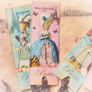 May include: Three vintage-style bookmarks with a Parisian theme. Each bookmark features a different image of a woman in a dress, with a pink, blue, and green color scheme. The bookmarks have the text "Paris" and "Marie Antoinette 1775" printed on them.