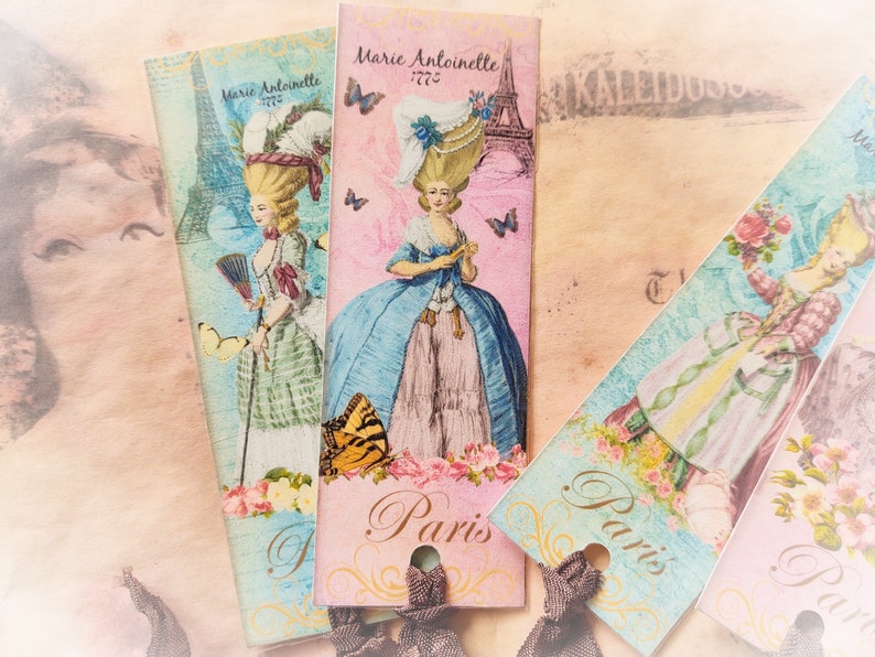 May include: Three vintage-style bookmarks with a Parisian theme. Each bookmark features a different image of a woman in a dress, with a pink, blue, and green color scheme. The bookmarks have the text "Paris" and "Marie Antoinette 1775" printed on them.