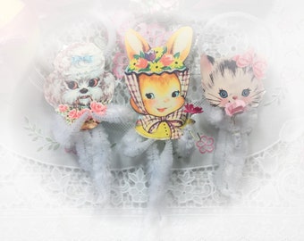 May include: Three vintage Easter decorations featuring a bunny, a poodle, and a kitten. Each figure is made of paper and has a white fluffy tail. The figures are arranged on a white lace tablecloth.