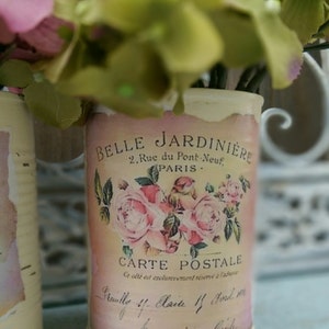 Op de afbeelding: Twee witte metalen blikken met bloemenmotieven en tekst. De tekst op de blik staat "Belle Jardiniere, 2 Rue du Pont Neuf, Paris, Carte Postale".