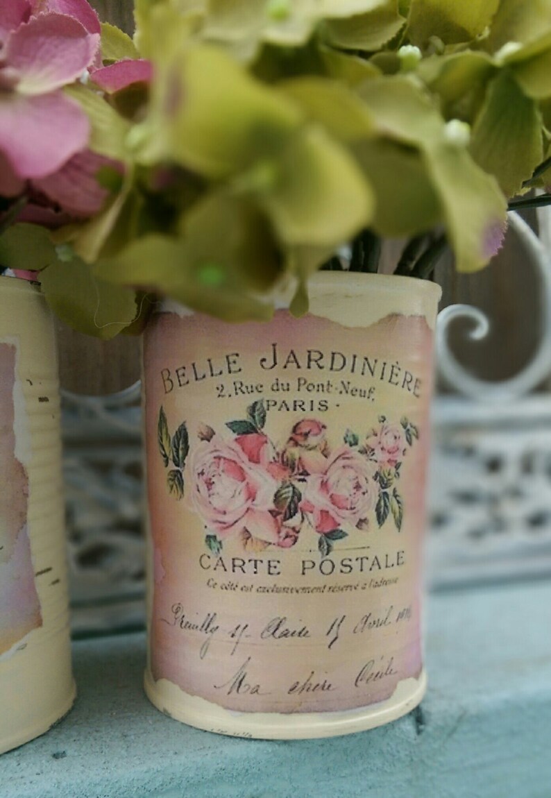 Op de afbeelding: Twee witte metalen blikken met bloemenmotieven en tekst. De tekst op de blik staat "Belle Jardiniere, 2 Rue du Pont Neuf, Paris, Carte Postale".