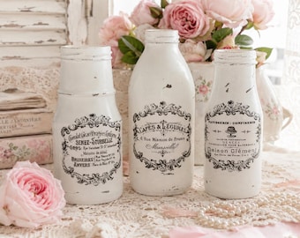 May include: Three white glass bottles with distressed paint finishes and black printed French typography labels. Each bottle features a unique decorative frame design with text including Senez-Sturbelle, Cafés & Légumes, and Maison Clément.