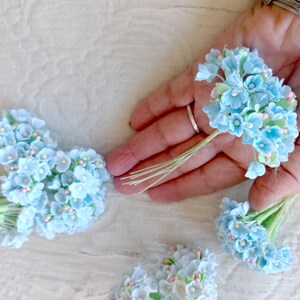 Pu&ograve; includere: Diversi piccoli mazzetti di fiori artificiali azzurri con minuscoli centri rosa e steli in filo metallico verde. Ogni mazzetto &egrave; composto da molteplici fiori in miniatura disposti in forma arrotondata, tenuti insieme da sottili steli flessibili.