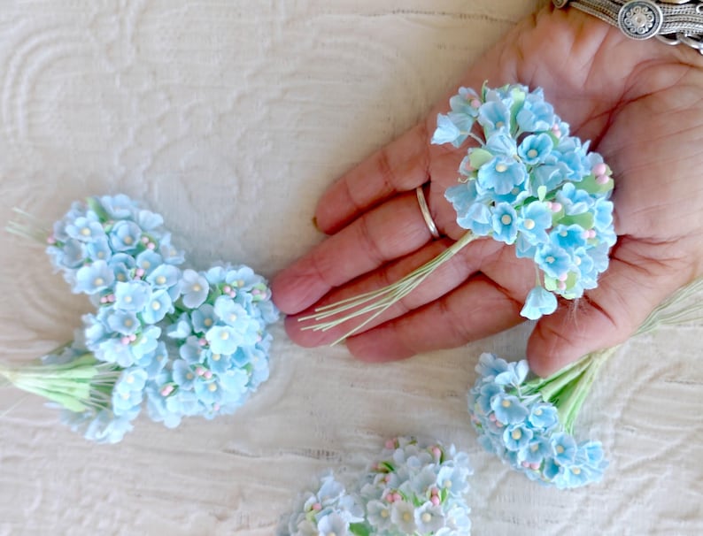 Pu&ograve; includere: Diversi piccoli mazzetti di fiori artificiali azzurri con minuscoli centri rosa e steli in filo metallico verde. Ogni mazzetto &egrave; composto da molteplici fiori in miniatura disposti in forma arrotondata, tenuti insieme da sottili steli flessibili.