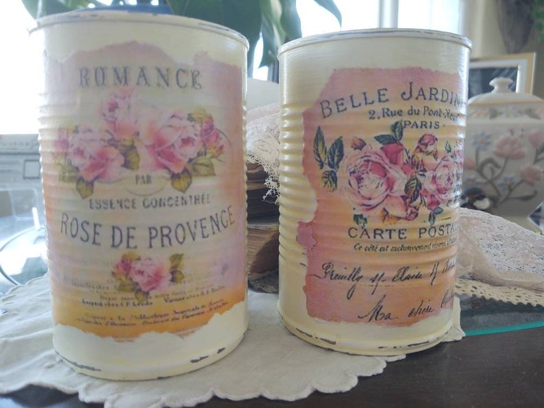 Op de afbeelding: Twee vintage-stijl metalen blikken met bloemmotieven en Franse tekst. De blik aan de linkerkant zegt "Romance" en "Rose de Provence". De blik aan de rechterkant zegt "Belle Jardin" en "Carte Postale".