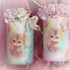 May include: Two vintage-style metal cans decorated with a pastel pink and blue background and a kitten image. The cans are filled with pink and white flowers and have a white lace ribbon tied around the top.