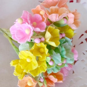 May include: A bouquet of pastel-colored artificial flowers. The arrangement features blossoms in shades of pink, yellow, and orange, with green leaves and small pink bead accents. The flowers are clustered together, creating a vibrant and delicate display.