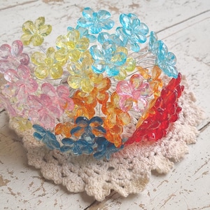 May include: A colorful floral hair accessory featuring translucent flowers in shades of pink, yellow, orange, blue, and red. The flowers are arranged on thin silver wires and rest on a cream-colored crocheted doily.