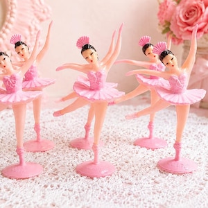 May include: A set of five plastic ballerina figurines, each wearing a pink tutu and a matching pink headpiece. The dancers are posed in various ballet positions, standing on circular pink bases. The figures have painted facial features.