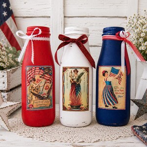 May include: Three decorative glass bottles painted red, white, and blue, with patriotic labels and ribbon accents. The bottles are decorated with vintage-style images and text, perfect for Fourth of July celebrations.