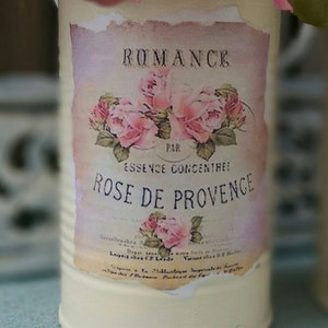 Op de afbeelding: Een vintage metalen container met een label waarop staat "Romance Par Essence Concentree Rose de Provence". Het label heeft roze rozen en groene bladeren. De container is wit geverfd met een verweerde afwerking.
