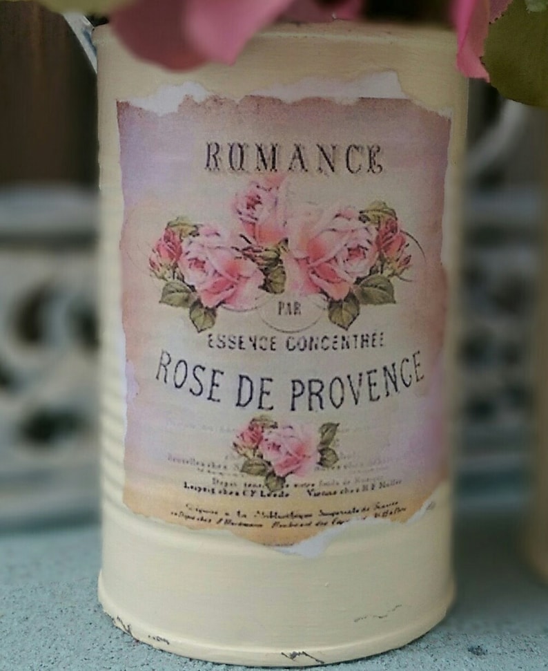 Op de afbeelding: Een vintage metalen container met een label waarop staat "Romance Par Essence Concentree Rose de Provence". Het label heeft roze rozen en groene bladeren. De container is wit geverfd met een verweerde afwerking.