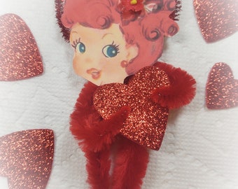 May include: A red glitter heart with a red fuzzy trim, a red glitter heart with a red fuzzy trim, and a red glitter heart with a red fuzzy trim. A paper cutout of a woman with red hair and a red flower in her hair. The woman is holding a red glitter heart with a red fuzzy trim.