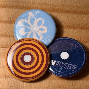 May include: Three round buttons with pop music designs. One is light blue with a flower and "Britney Spears" text. Another is orange and maroon with concentric circles and "No Strings Attached" text. The third is dark blue with "NSYNC" text.