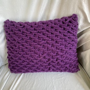 May include: Purple crocheted pillow with a textured pattern. The pillow is on a white surface.