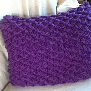 May include: Purple crocheted pillow with a textured, scale-like pattern. The pillow is sitting on a white couch.