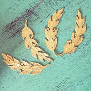 Op de afbeelding: Vier gouden metalen lauwerkransen met gedetailleerde bladontwerpen. De kransen zijn in een halve cirkel op een turquoise achtergrond gerangschikt.