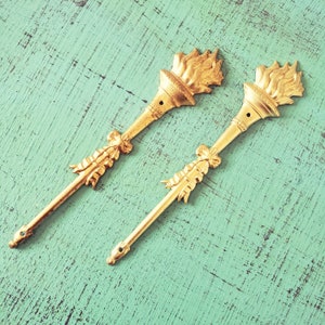 Peut inclure: Deux appliques murales décoratives en métal doré avec un motif de torche et un détail de nœud. Les appliques sont sur un fond bleu sarcelle.