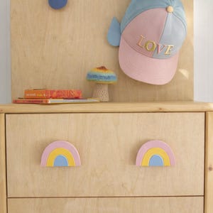 Peut inclure: Une commode en bois clair avec des poignées en forme d'arc-en-ciel. Une casquette de baseball bleue et rose avec le mot "LOVE" est suspendue au-dessus. Un petit champignon crocheté et des livres sont posés dessus.