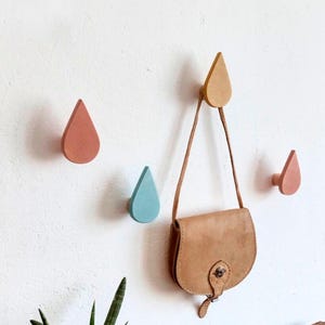 Peut inclure: Quatre crochets muraux en bois en forme de gouttes de pluie de différentes couleurs : jaune, rose, bleu et marron clair. Un sac à main en cuir marron est accroché au crochet jaune.