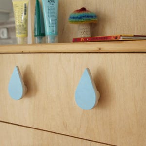 Peut inclure: Deux poignées de tiroir en forme de goutte d'eau bleu clair sur une armoire en bois clair. Les poignées ont une base arrondie et un devant plat peint. Une étagère au-dessus contient des tubes de peinture, un champignon crocheté et un livre.