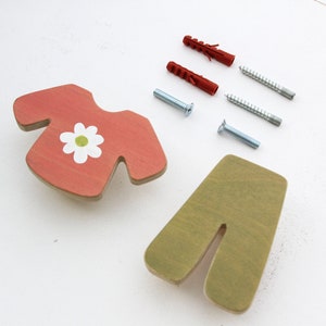 Peut inclure: Deux crochets muraux en bois en forme de chemise et de pantalon. La chemise est rouge avec une fleur blanche et un centre vert. Le pantalon est vert. Les crochets sont livrés avec des vis et des chevilles pour l'installation.