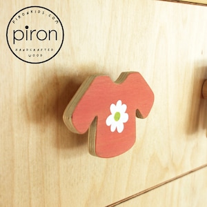 May include: A red wooden drawer pull shaped like a t-shirt with a white flower on it. The pull is attached to a light wood surface. The text "piron handcrafted wood" is visible in a circle in the upper left corner of the image.