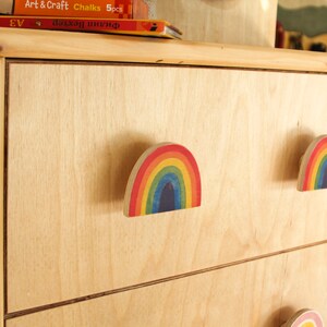 Könnte beinhalten: Eine Kommode aus Holz mit drei Schubladen. Jede Schublade hat einen regenbogenfarbenen Holzgriff. Die Kommode ist aus hellem Holz.