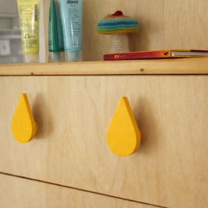 Op de afbeelding: Houten kast met twee gele, druppelvormige ladeknopen. Bovenop staat een plank met tubes lotion, een gehaakte paddenstoel en een klein boek. Eenvoudig en modern ontwerp.