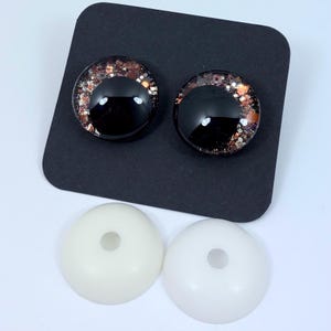 May include: Two black, glossy doll eyes with glitter accents in shades of copper and gold, set on a black background. Below are two white, round plastic pieces with a hole in the center. These are likely the backs of the eyes.